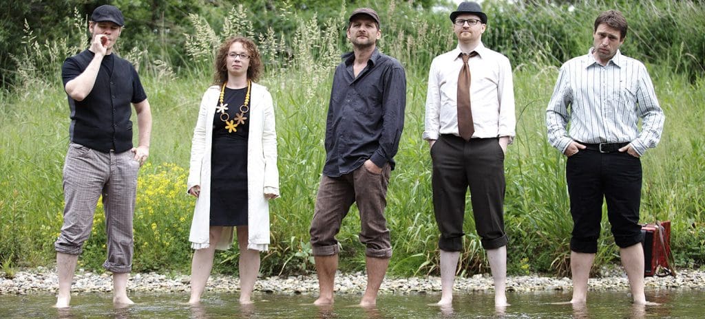 Die fünfköpfige Band mit aufgestickten Hosen in einem Bach oder Flussbett. Im Hintergrund ist Schilf und grüne Vegetation zu sehen.