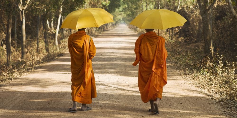 Zwei Mönche in orangen Gewändern und gelben Sonnenschirmen alleine auf einem Weg