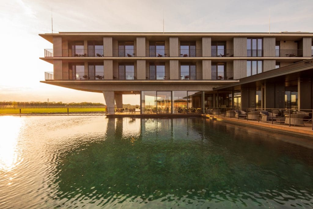 The Resort mit niedrig stehender Sonne und Reflexionen am Wasser davor. Im Hintergrund sind Weinberge angedeutet.