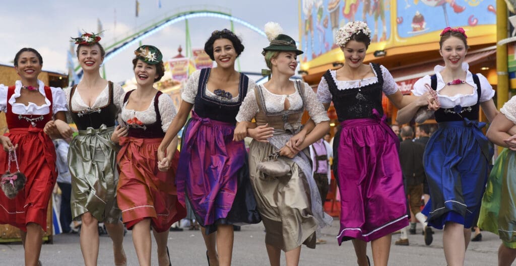 Frauen gehen eingehängt und tragen bunte Dirndl. Im Hintergrund ist ein Vergnügungspark zu sehen.