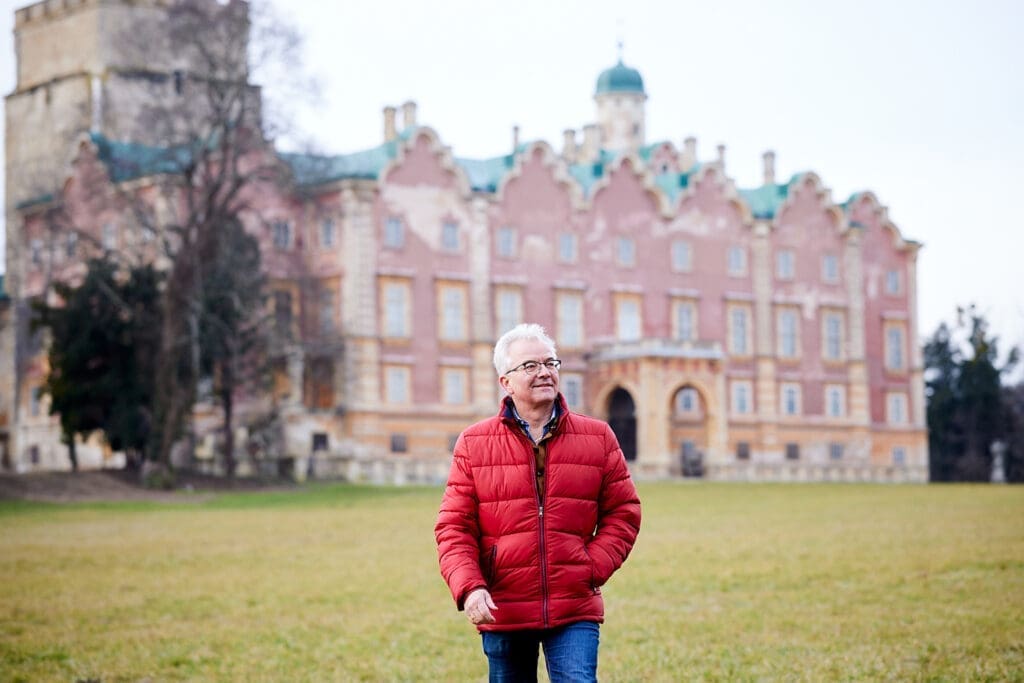 Bürgermeister Gerhard Weil in roter Steppjacke vor dem Schloss Prugg im Harrachpark. Bäume und Wiese haben eine herbstliche Färbung.