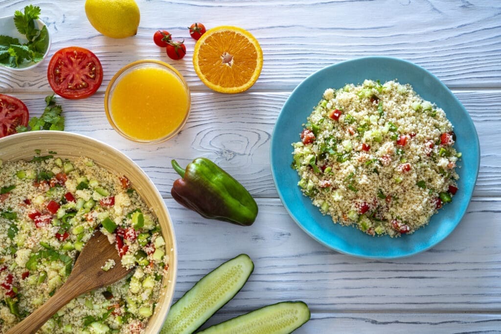 Couscous-Salat mit Radieschen und Gurke auf einem azurblauen Teller. Daneben Couscous-Salt in einer Holzschüssel mit einem Holzlöffel. Auf dem Tisch befindet sich weiters Orange, Orangensaft, Tomate, Paprika und Gurke appetitlich dekoriert.