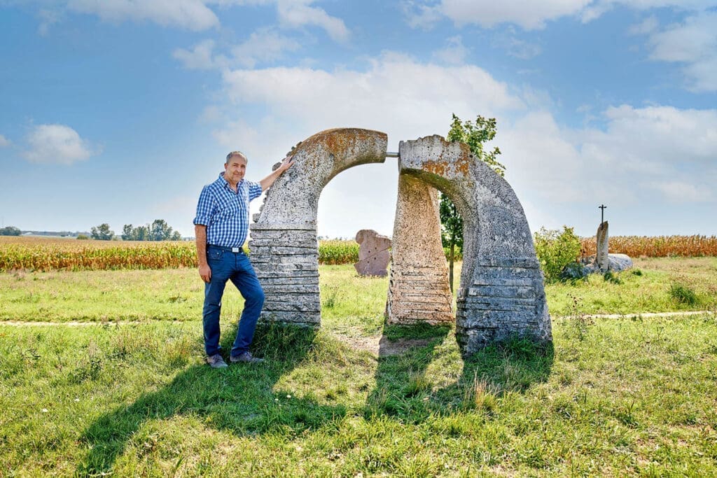 Bürgermeister Gerhard Bachmann Skulpturenpark an eine Steinfigur gelehnt. Im Hintergrund sieht man weitere Figuren, Felder und den blauen Himmel.