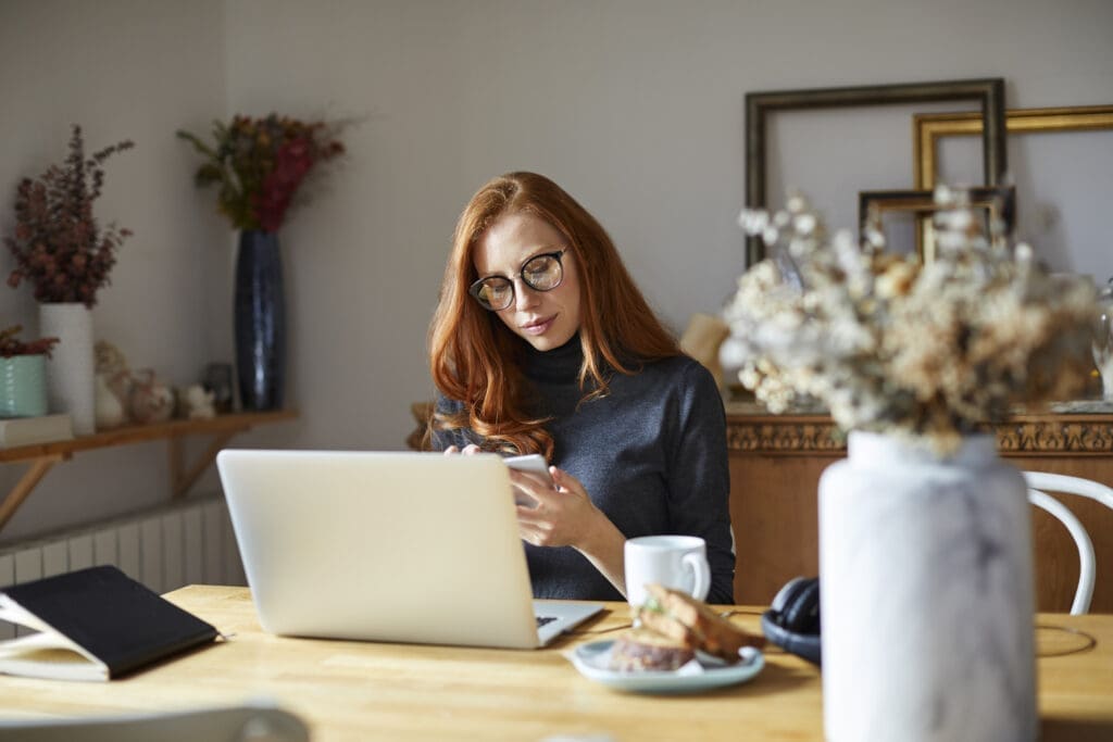 Frau sitzt mit Handy am Tisch, vor ihr ein Laptop