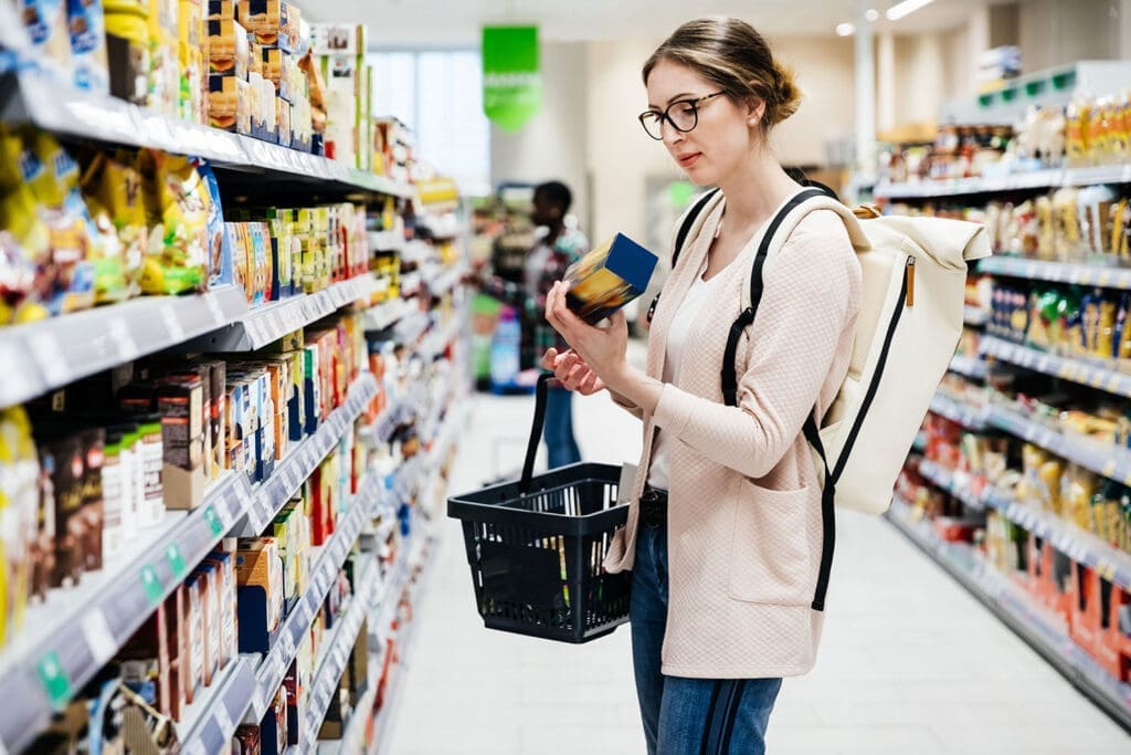 Frau mit Brille, Knoten und rosaroter Weste in Supermarkt mit Korb in der einen und Produkt in der anderen Hand.