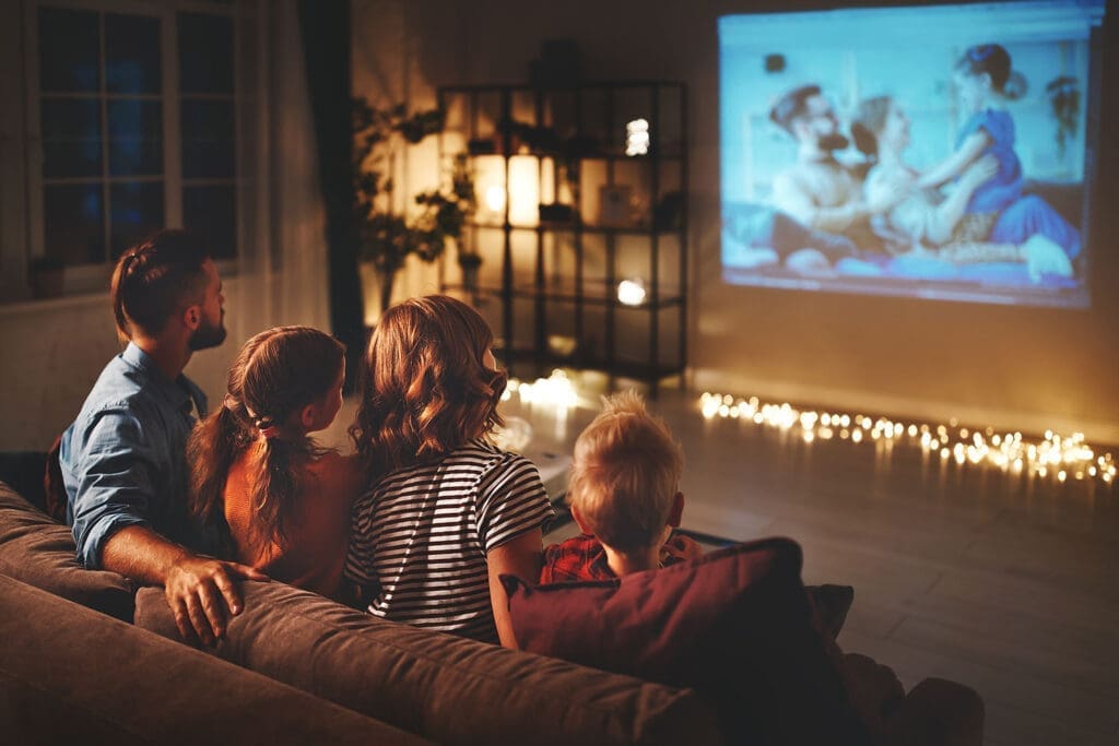 Vater, Mutter, Tochter und Sohn sehen abends auf der Couch über eine Beamer-Leinwand fern. Im Hintergrund sanftes, indirektes Licht und Lichterketten am Boden.