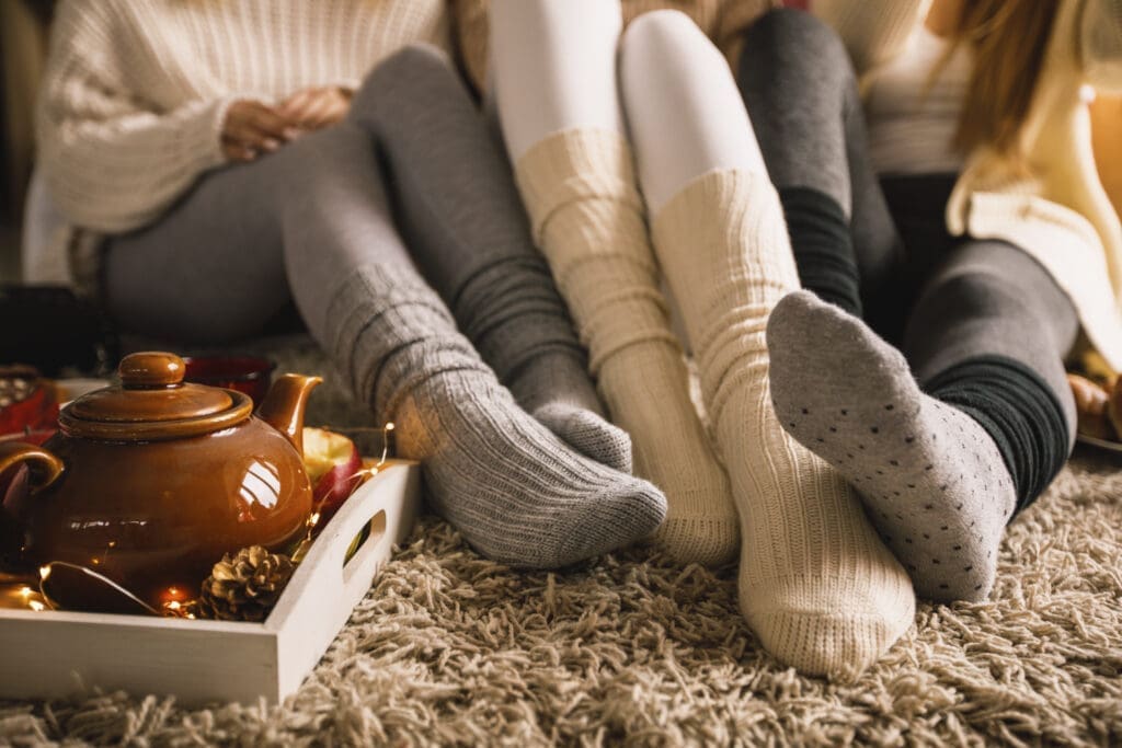 Drei Fußpaare mit kuscheligen Socken auf einem Teppich mit einer Teekanne daneben.