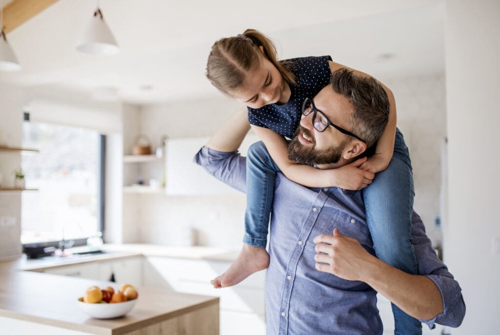 Gentle Parenting: Vater mit Tochter auf den Schultern in weißer Küche.