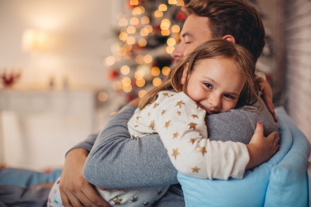 Vater hält Mädchen auf dem Arm, hinter den beiden steht ein Christbaum