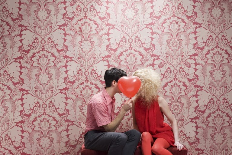 Mann mit weiß-kariertem Hemd, schwarzer Hose und schwarzem Hemd und Frau in rotem Kleid und roten Leggings auf rotem Polstermöbel vor einer gemusterten Tapete in creme und rot bei ihrem Date. Ein roter Herzluftballon verdeckt ihre Gesichter.