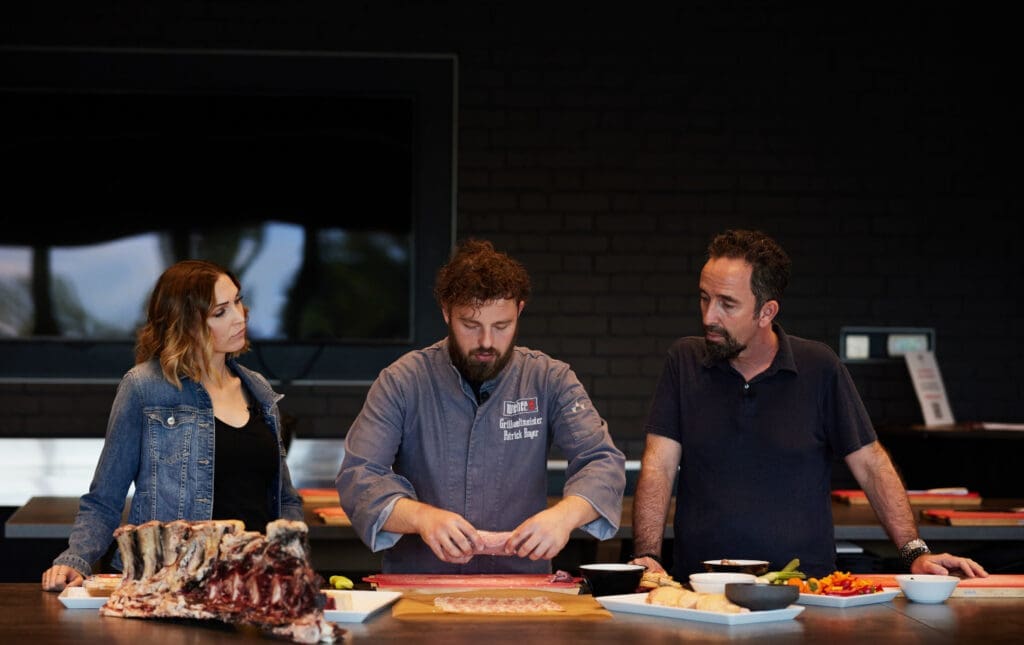 Grillweltmeister Patrick Bayer in der Mitte zeigt Redaktionsleiterin Bernadette Strobl und Verlagsleiter Christoph Berndl, wie Fleisch und Gemüse für den Grill vorbereitet werden.
