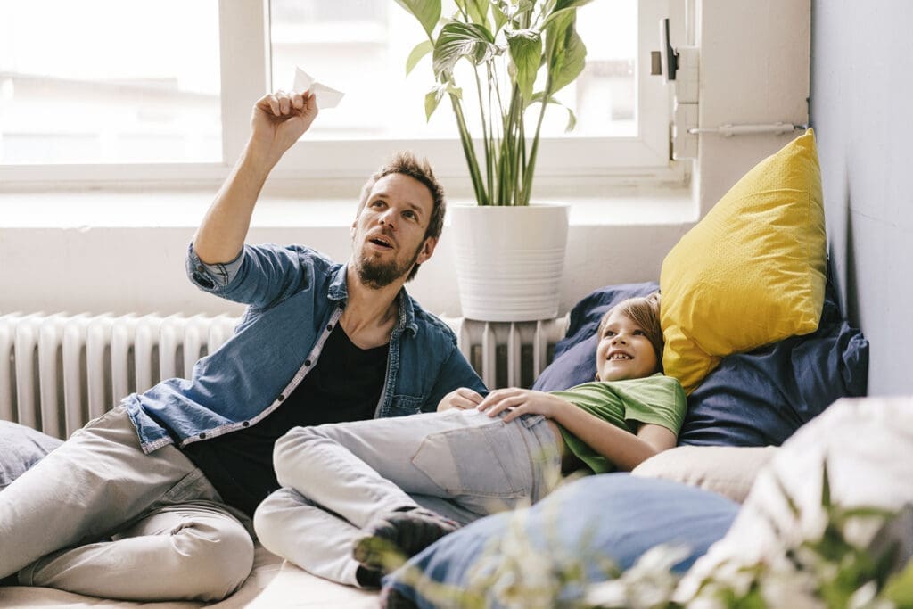 Mann mit Papierflieger in der Hand in liegender Position auf Sofa mit vielen Kissen neben Jungen.