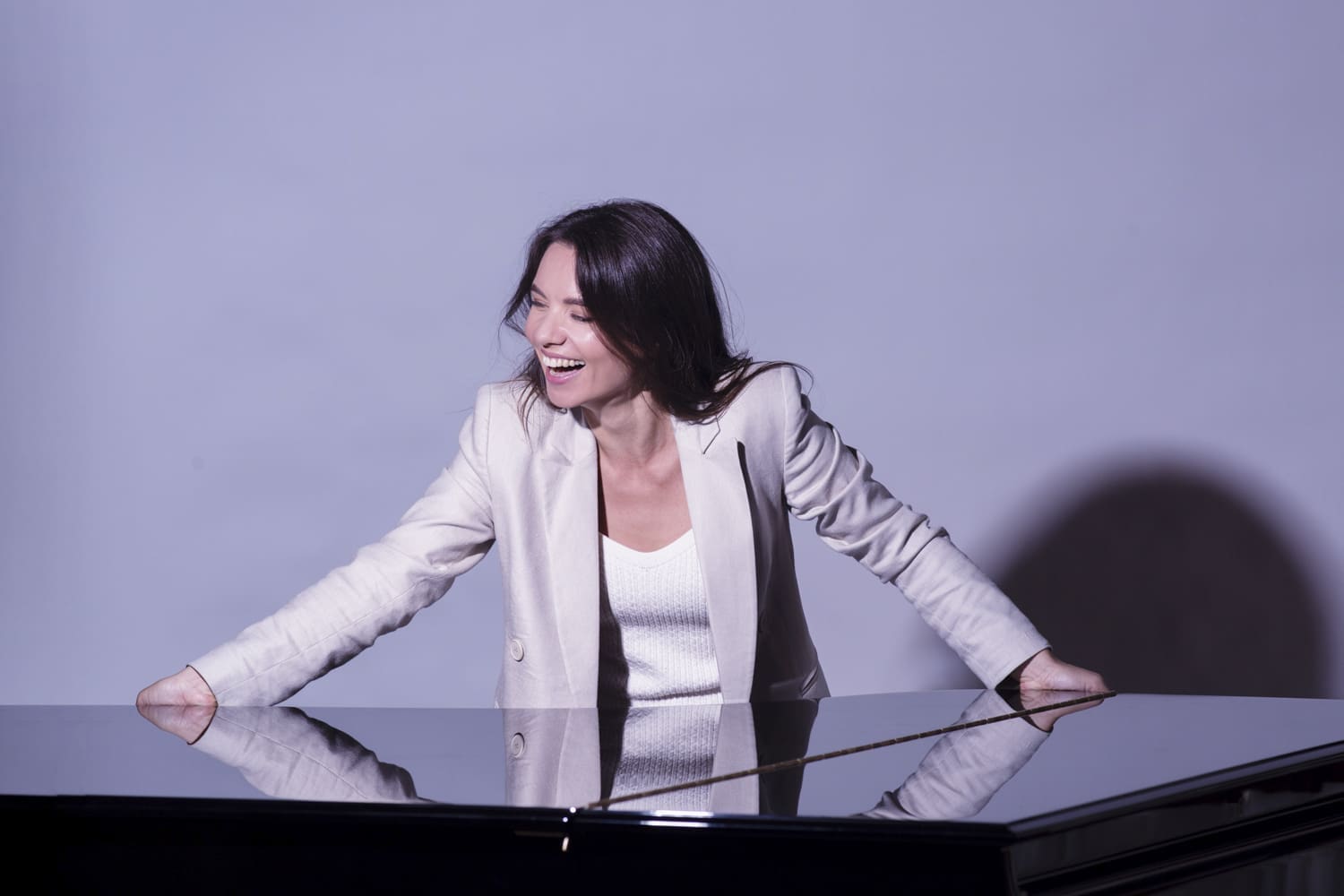Kateryna Titova in weißem Shirt und grauem Blazer lacht mit beiden Händen auf ein Klavier gestützt