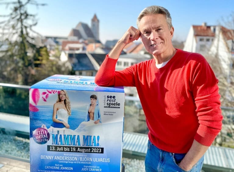 Alfons Haider lächelnd mit aufgestützter Hand auf einem Werbewürfel von Mamma Mia! in einem kirschroten Pullover. Im Hintergrund sieht man verschwommen die Umrisse einer Gemeinde.