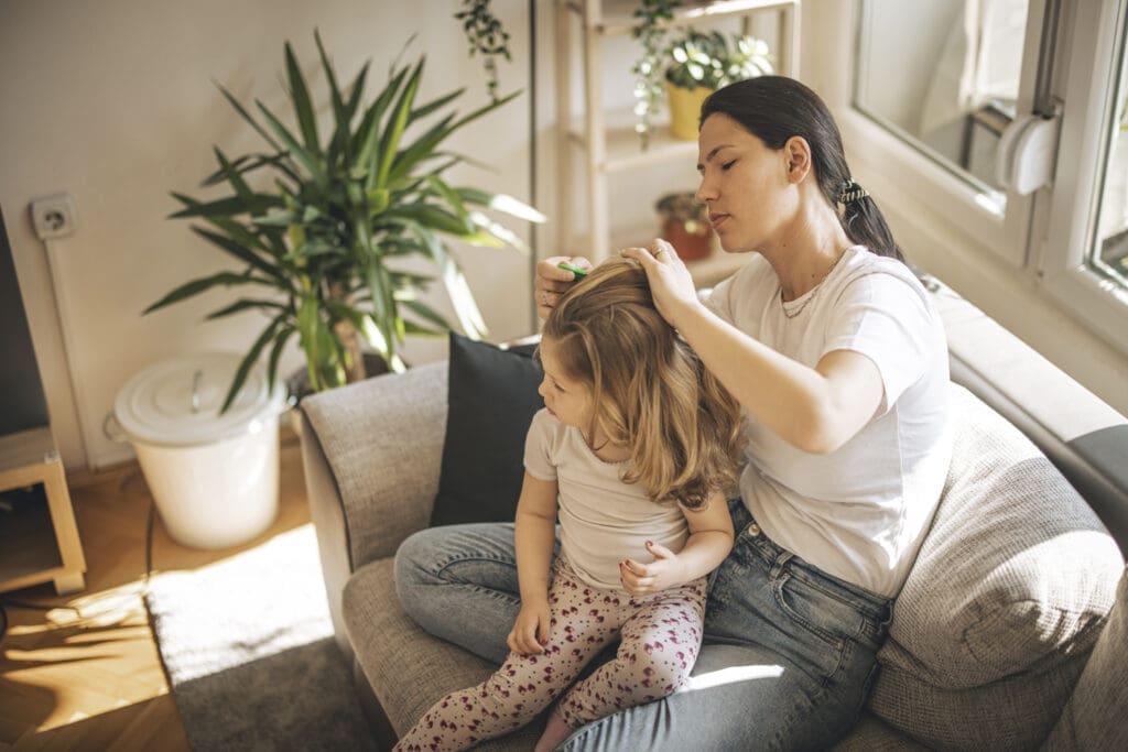 Mutter sitzt mit Tochter auf der Couch und sucht den Kopf des Kindes nach Läusen ab.