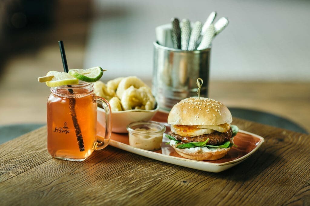 Burger von Le Berger mit Limonade auf Holztisch. Im Hintergrund Besteck in einem Metallgefäss.