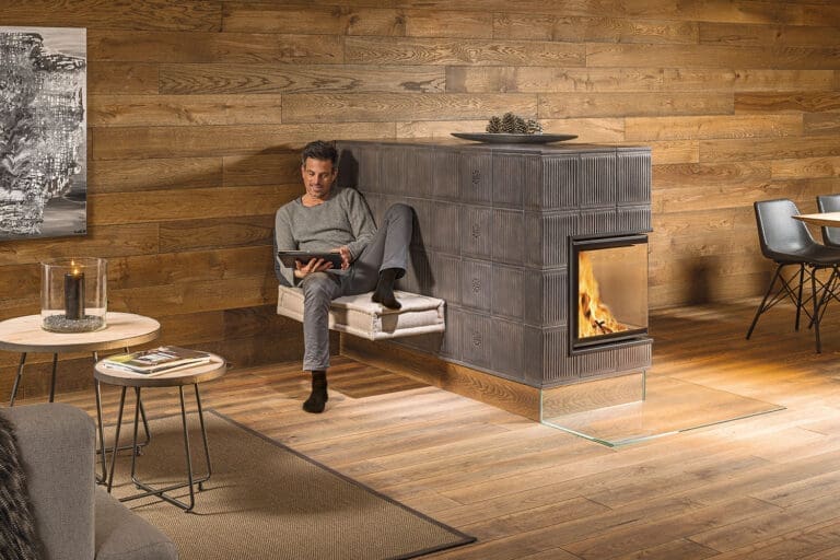 Mann in entspannter Haltung mit Tablet auf dem Schoß auf einer Ofenbank neben einem offenen Kachelofen. Wand und Boden sind aus dunklem Holz.