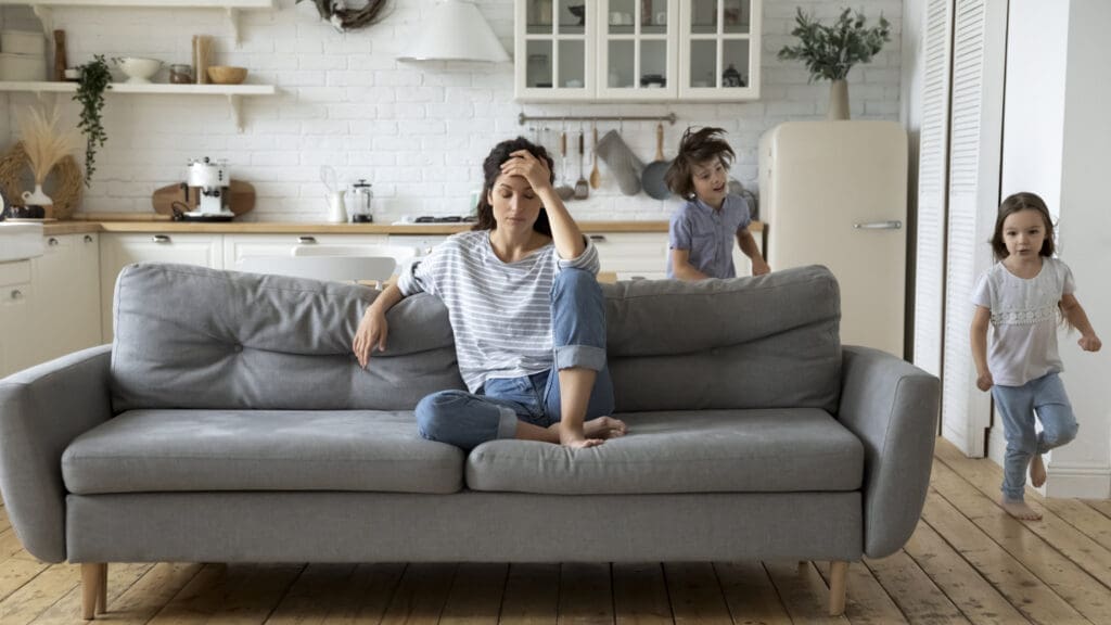 Frau sitzt gestresst auf Couch, um sie herum laufen zwei Kinder