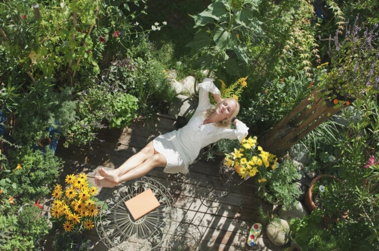 Frau liegt in einem Stuhl in einem Garten aus grünen Pflanzen mit den Füßen auf dem Tisch. Auf dem Tisch liegt ein Buch. Sie hat die Hände hinter dem Kopf verschränkt und lächelt mit geschlossenen Augen.
