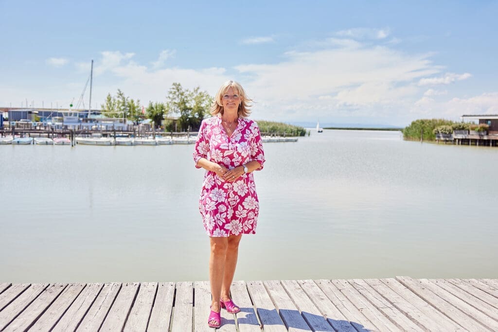 Bürgermeisterin Elisabeth Böhm im wunderschönen Hafen ihrer Gemeinde. Im Hintergrund sieht man Stege, Boote, Schilf und einen azurblauen Himmel mit einigen weißen Wolken.