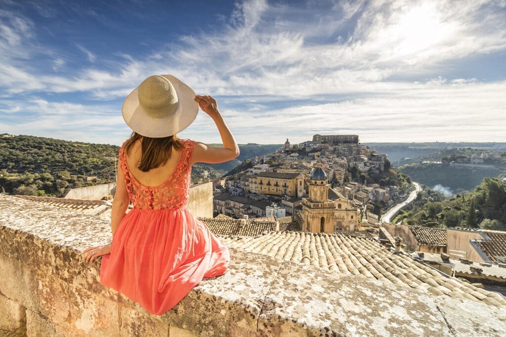 Frau mit Sonnenhut sitzt auf einer Steinmauer in der sizilianischen Stadt Ragusa mit Blick auf den Ort Ibla