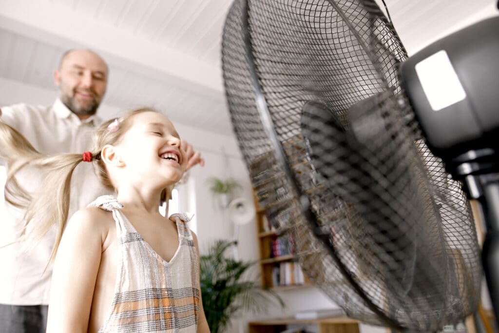 Kind steht vor Ventilator und genieß den Luftzug, hinter ihr steht ihr Vater