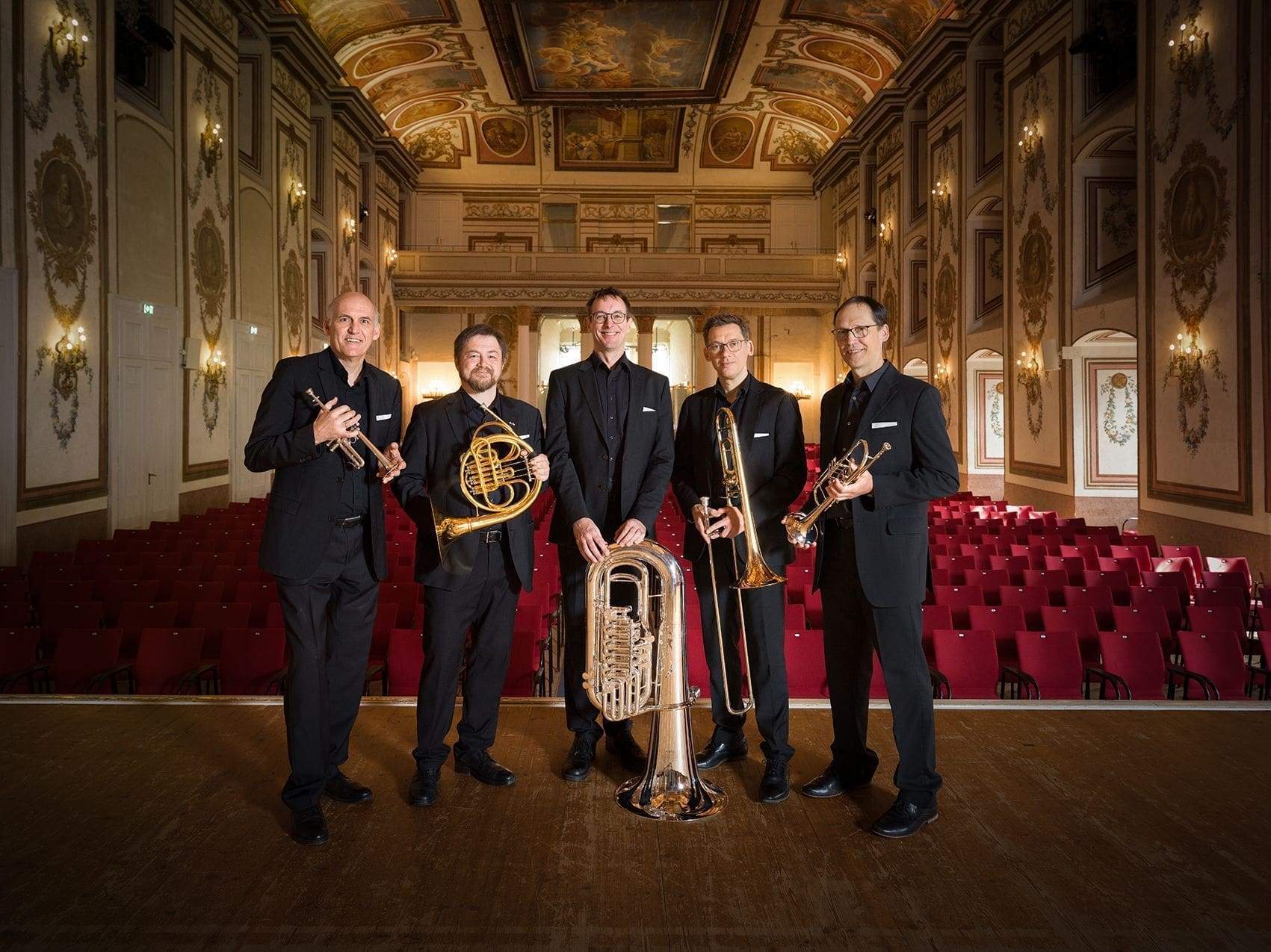 Vier Blasmusiker mit ihren Instrumenten auf einer Bühne in einem Prunksaal voller roter Sessel