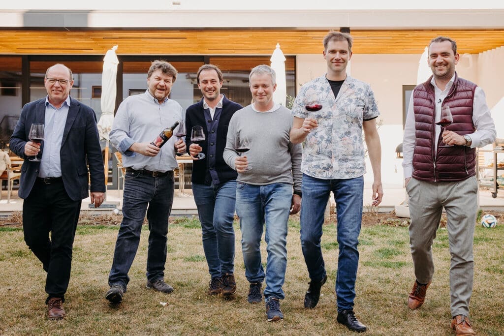 Die vier Winzer und Weintourismus-Obmann Herbert Oschep sowie Weinbaupräsident Andreas Liegegeld mit einem Glas Rotwein in der Han auf einer Wiese.