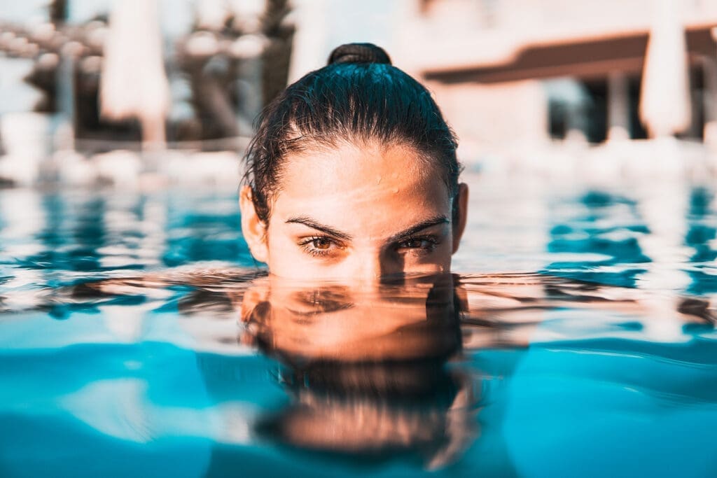 Frau halb im Wasser. Sie hat einen Zopf und nasse Haare. Aus dem Wasser heraus schaut die obere Hälfte ihrer Nase, Augen und Stirn. Das Wasser ist azurblau und glasklar.