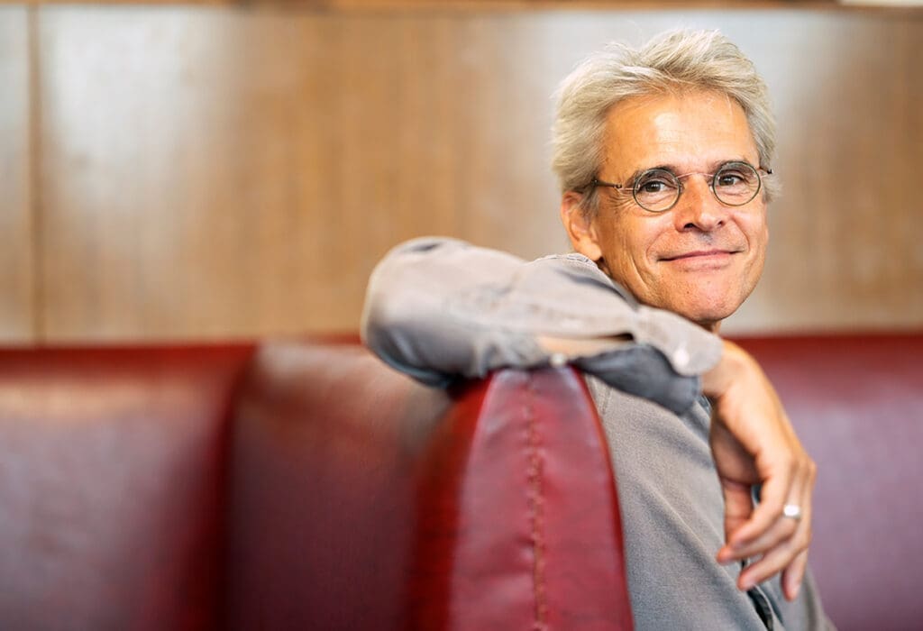 Thomas Brezina in grauem Hemd mit runder Brille in nonchalanter Pose auf einer roten Lederbank.