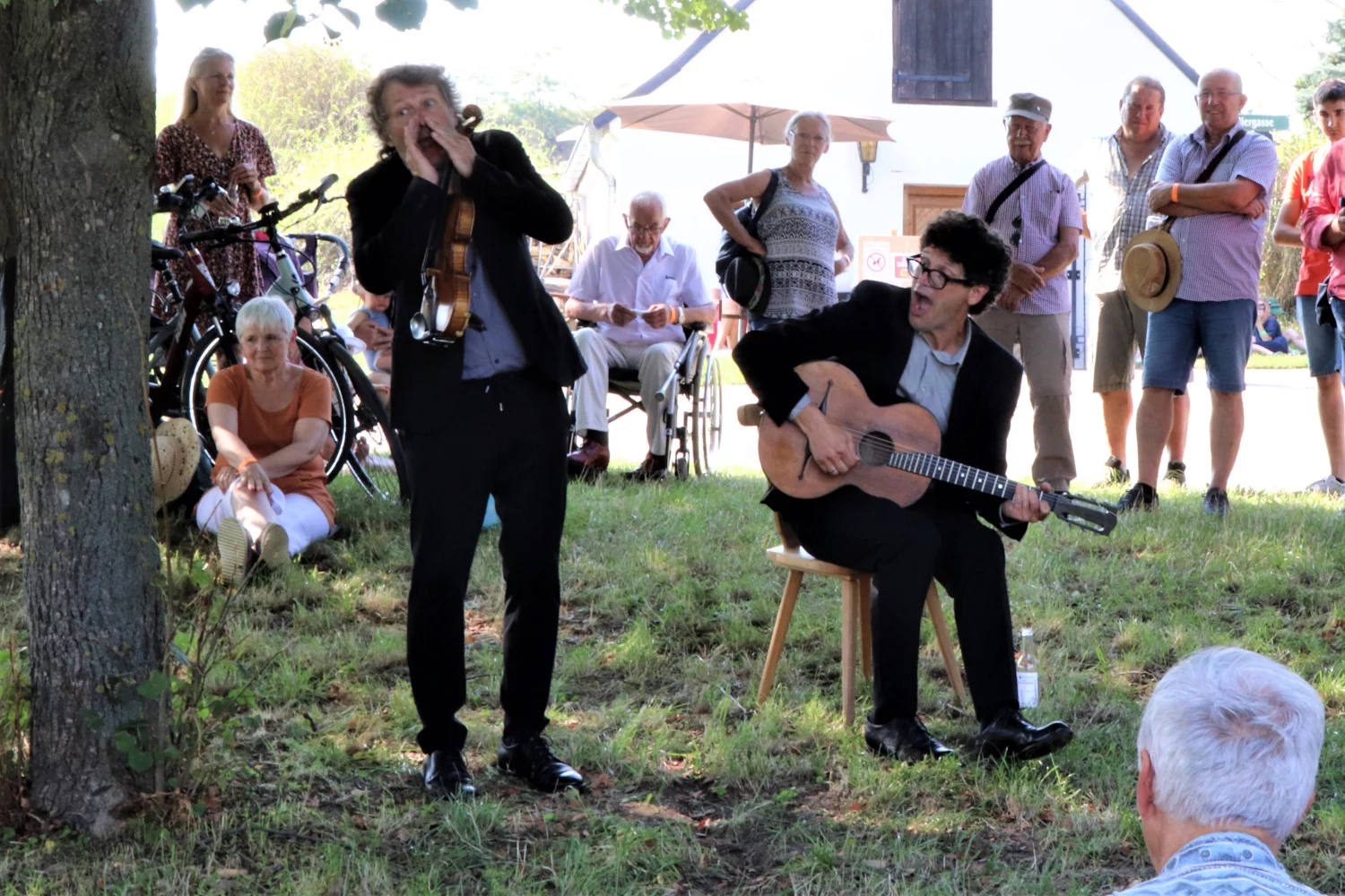 Zwei Musiker stehen auf einer Wiese im Freien und spielen ihre Instrumente, rings um sie herum steht bzw. sitzt das Publikum