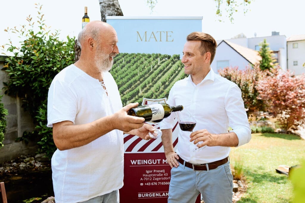 Winzer Mate in kurzärmeligen T-Shirt schenkt Bürgermeister Ivan Grujic Rotwein in Glas ein.