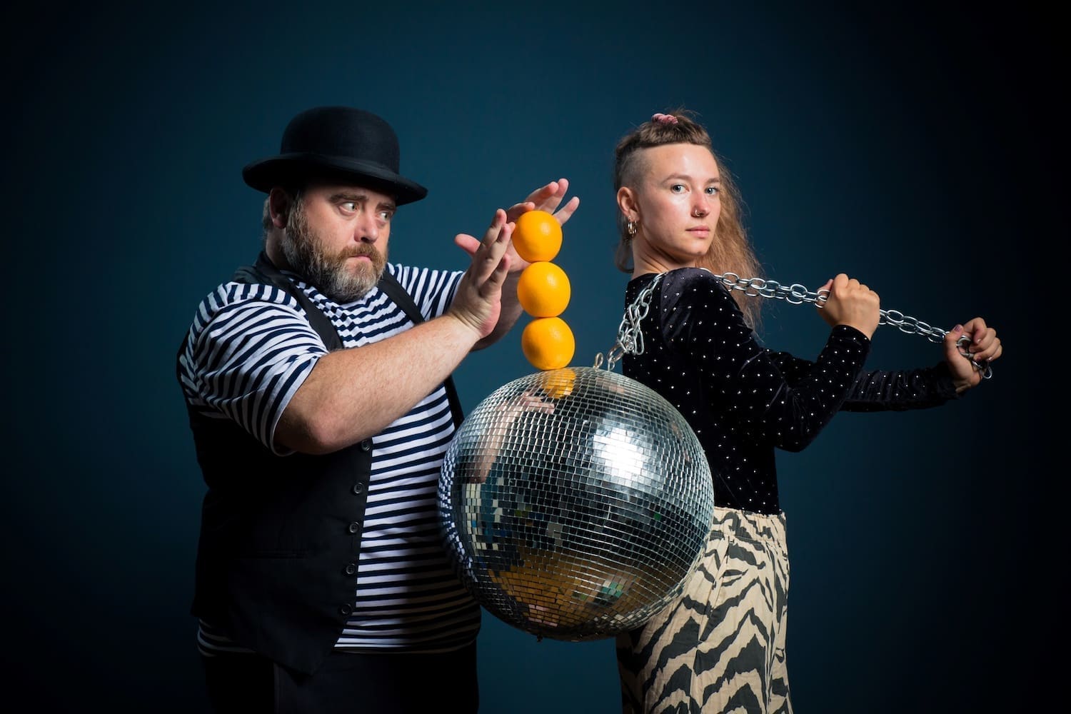 Eine Frau trägt eine Discokugel an einer Kette über der Schulter, ein Mann mit Hut stapelt Mandarinen auf Kugel übereinander. („Obstacles in our Sky“) © Tom Weilguny