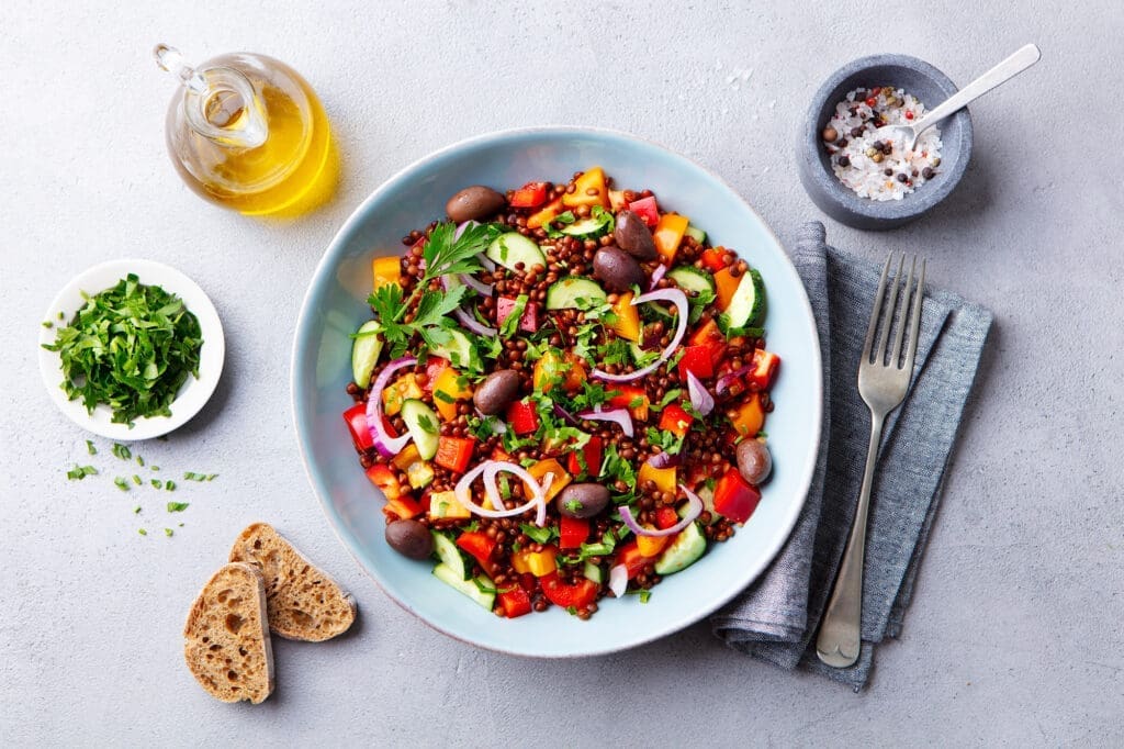 Linsensalat auf Teller vor grauem Hintergrund. Daneben grobkörniges Salz, Öl und Petersilie sowie Brot.