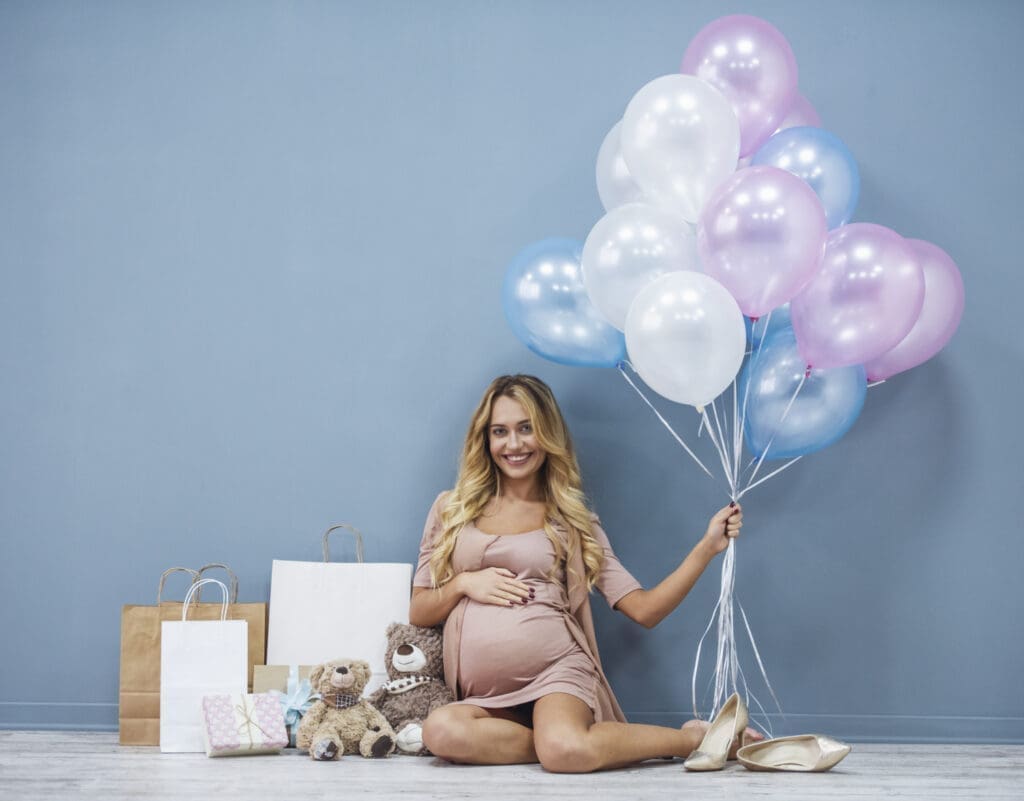 Babyparty: Schwangere Frau mit Geschenken und Luftballons in der Hand vor blauer Wand.