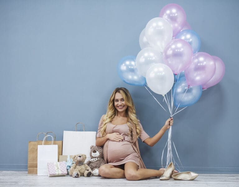 Babyparty: Schwangere Frau mit Geschenken und Luftballons in der Hand vor blauer Wand.