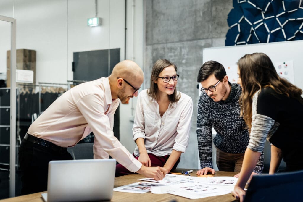 Vier-köpfiges Team arbeitet an einem Schreibtisch aus Holz mit Unterlagen und Laptop.