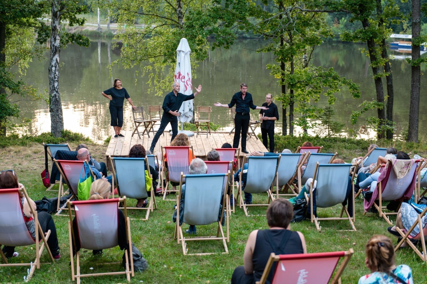 Publikum in Liegestühlen auf einer grünen Wiese vor einer Bühne im Freien, im Hintergrund ein See