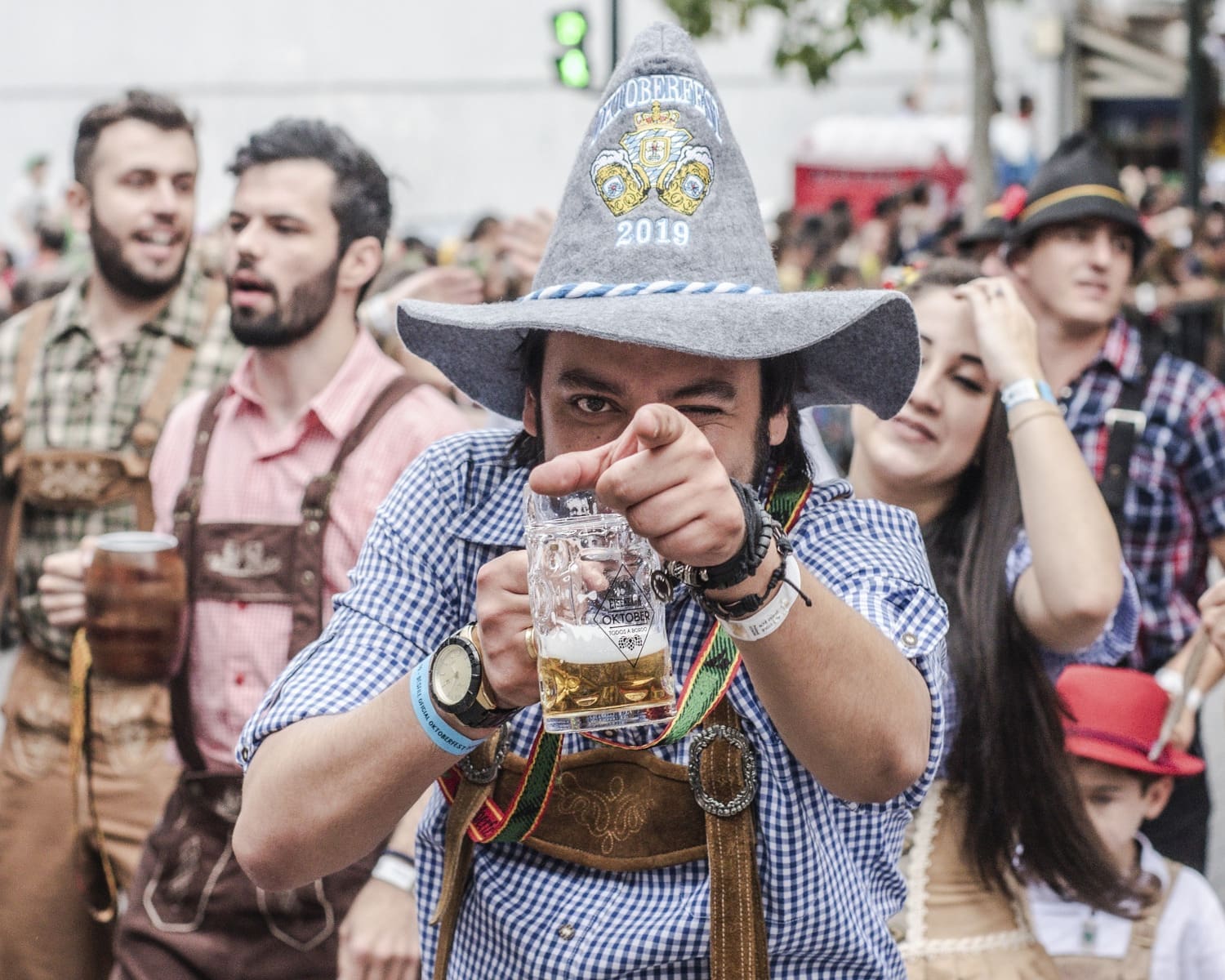 Mann in Tracht mit Filzhut deutet mit dem Zeigefinger seiner linken Hand in Richtung Kamera, in der rechten Hand hält er eine Maß Bier