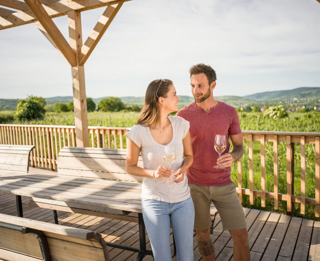 Frau und Mann stehen mit einem Glas Wein in der Hand auf einer Terasse/Aussichtsplattform aus Holz. Aufnahme von vorne