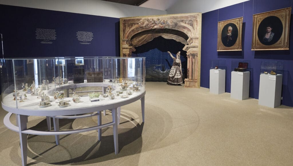 Ausstellungsansicht mit Vitrine und Gemälden im Hintergrund