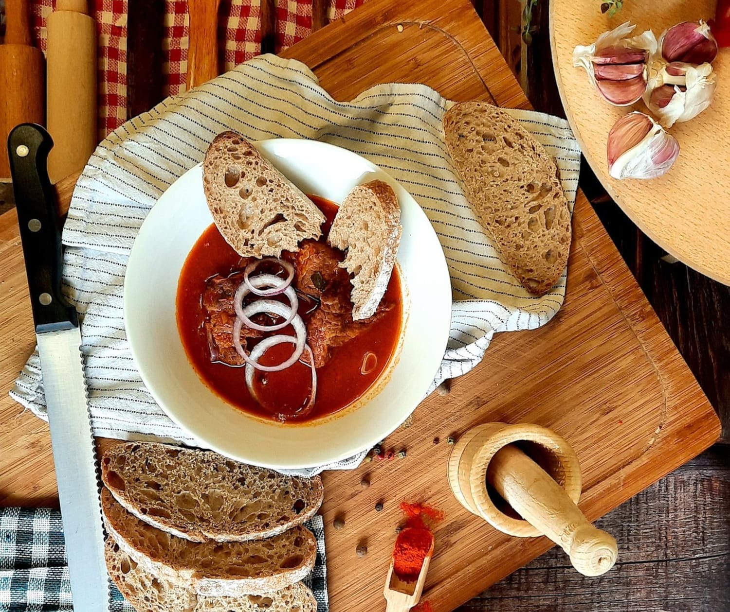 Gulasch in weißer Schüssel auf Holzbrett von Rohzutaten umringt